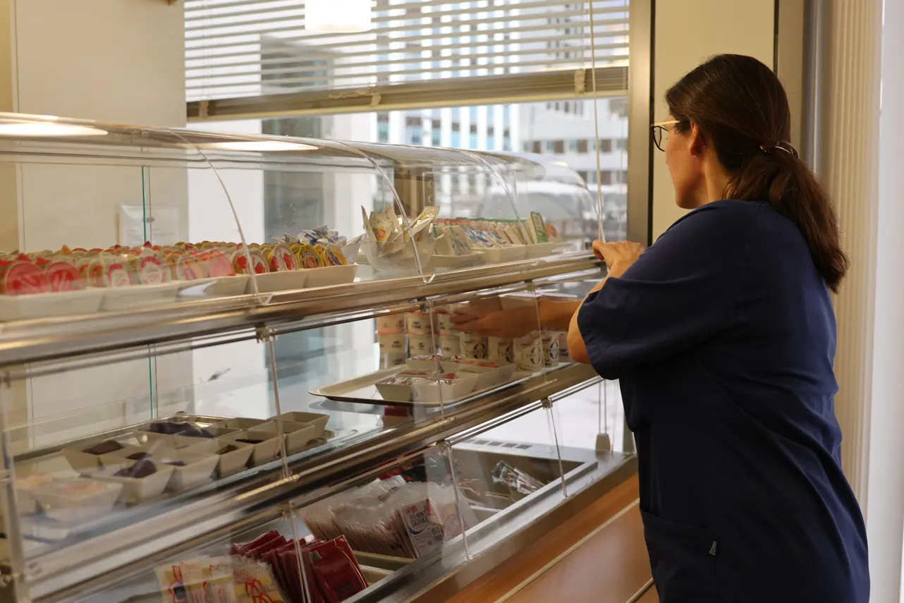 Ein person i arbeidsklede står ved ein sjølvbetent kantinedisk og fyller på med små yoghurtbeger. I disken ligg ulike porsjonspakkar med pålegg og tilbehøyr.